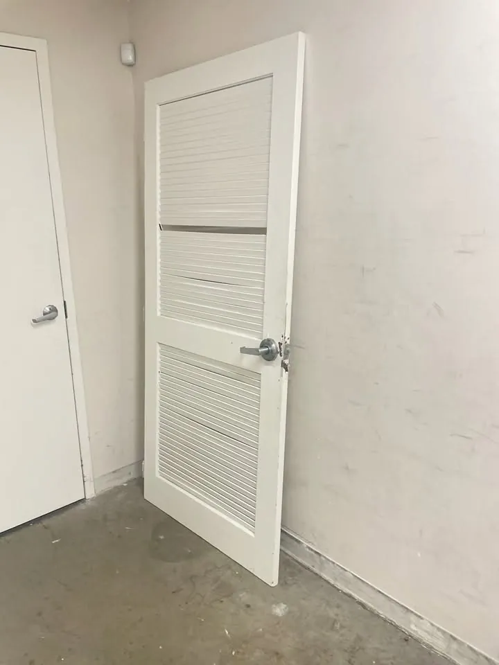 White louvred door with a damaged frame leaning against a stockroom wall
