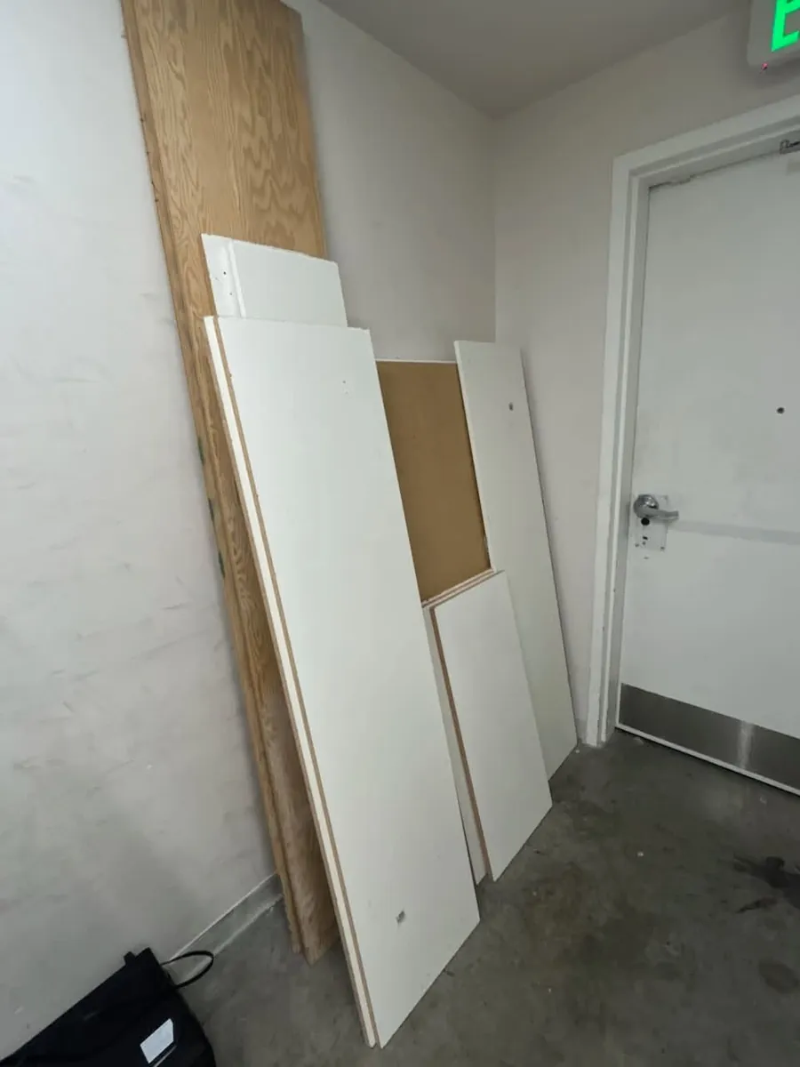 Plywood and white shelving panels propped against the wall of a store back room beside a fire door
