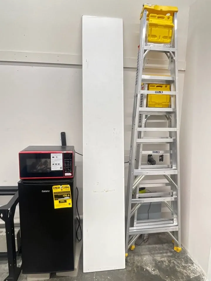 Long white shelf panel leaning against a store room wall between a mini fridge and a stepladder