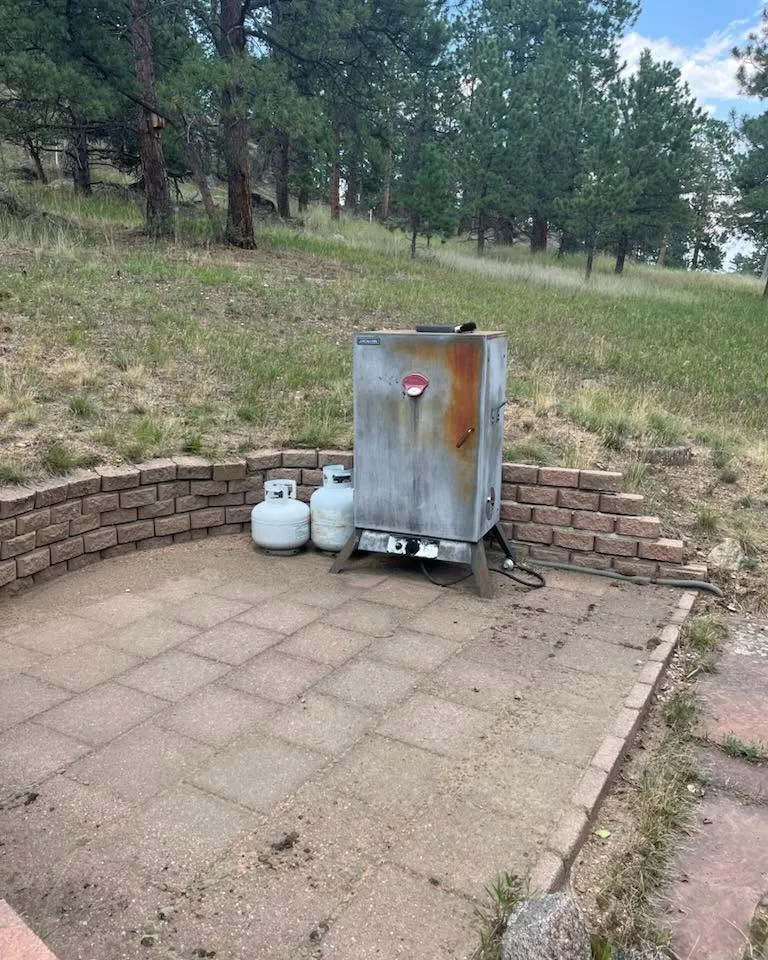 An old smoker and two propane tanks on a patio behind a stone wall, pine trees on the hill above, in Evergreen