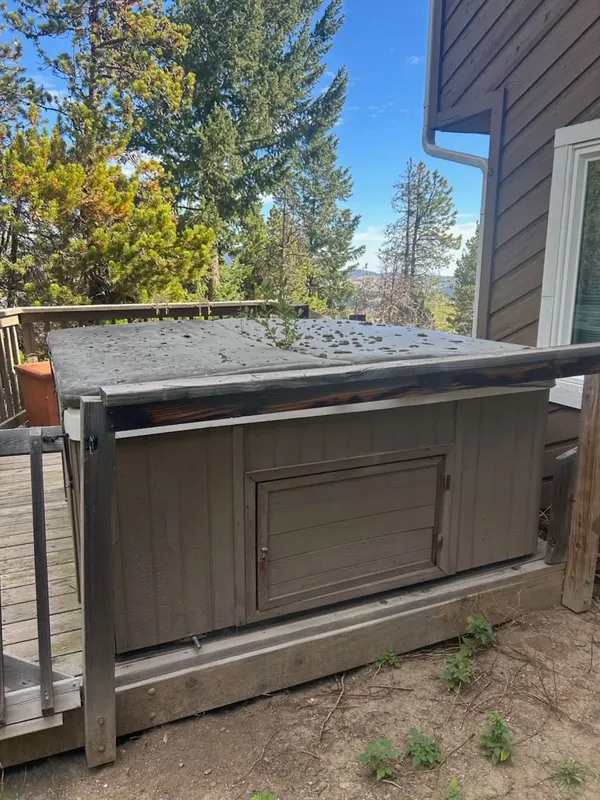 Old hot tub with a weathered cover sitting on a wooden deck beside a house, pine trees behind