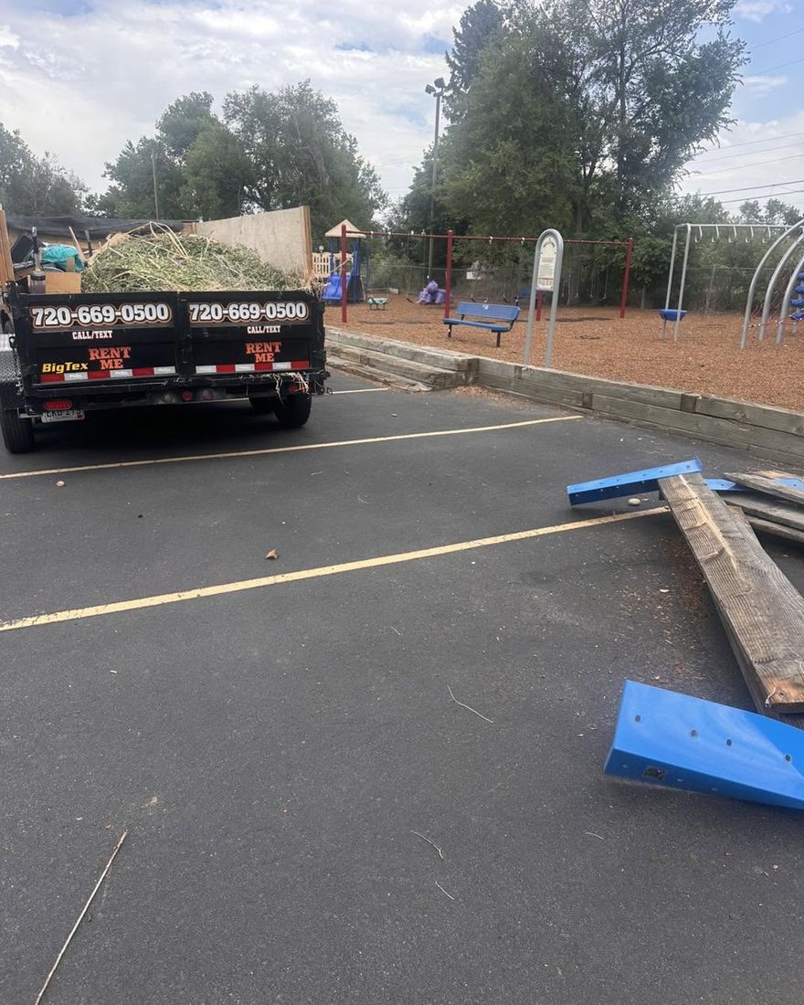 The Bob's Round Up trailer loaded with yard waste beside a Lakewood school playground, with old timbers and blue play equipment panels stacked on the lot