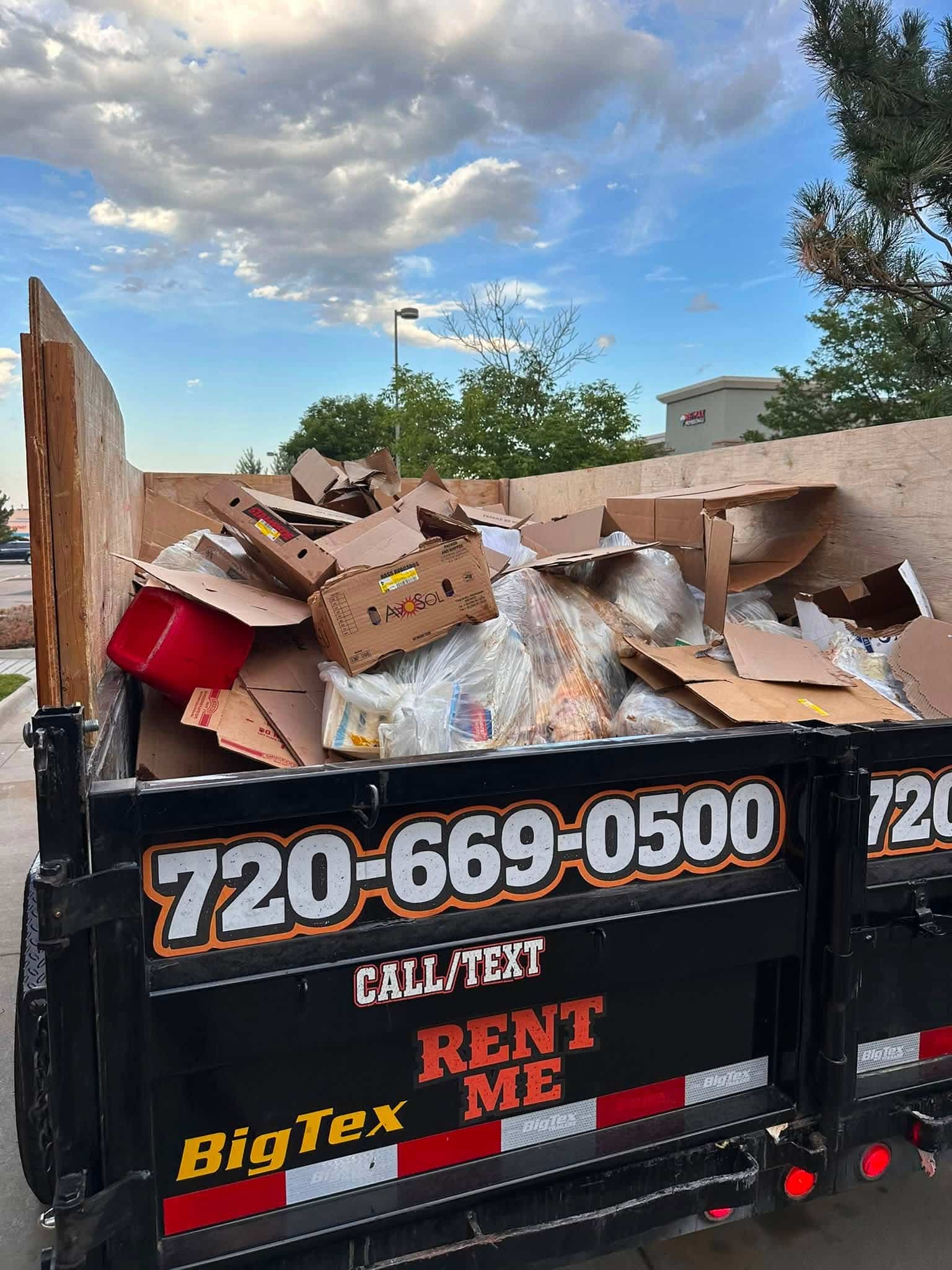 Black dump trailer loaded with flattened cardboard, clear bags of waste, and a red container